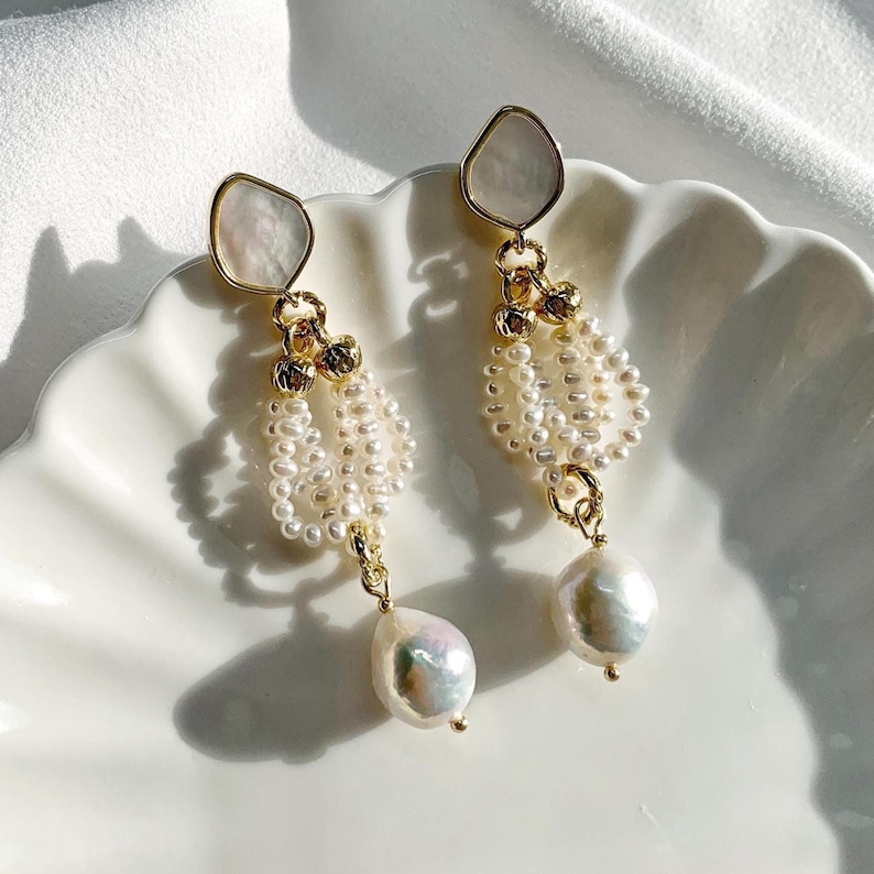 May include: A pair of gold-toned drop earrings featuring a top irregular-shaped white shell component, a central section composed of multiple strands of small white seed pearls, and a single large, lustrous white baroque pearl at the base.