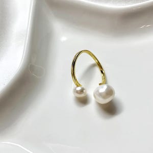 K&ouml;nnte beinhalten: Ein goldfarbener, offener Ohrring mit zwei wei&szlig;en Perlen. Eine Perle ist kleiner als die andere. Der Ohrring ist vor einem wei&szlig;en Hintergrund platziert.