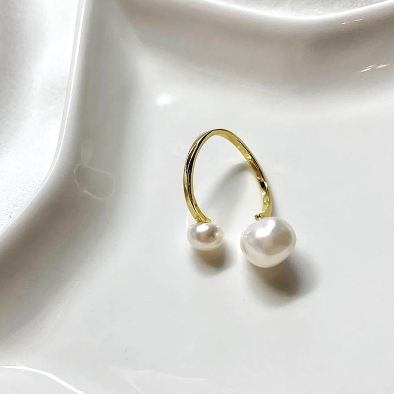 K&ouml;nnte beinhalten: Ein goldfarbener, offener Ohrring mit zwei wei&szlig;en Perlen. Eine Perle ist kleiner als die andere. Der Ohrring ist vor einem wei&szlig;en Hintergrund platziert.