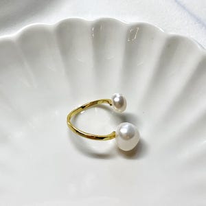 K&ouml;nnte beinhalten: Ein goldfarbener offener Ring mit zwei gro&szlig;en, wei&szlig;en Barockperlen. Der Ring hat ein schlichtes, elegantes Design, wobei die Perlen an den Enden des offenen Bandes positioniert sind. Der Ring liegt auf einem wei&szlig;en, wellenf&ouml;rmigen Hintergrund.