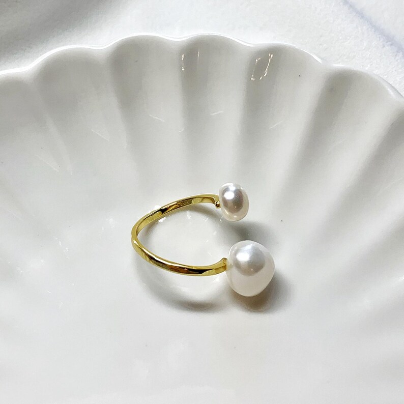 K&ouml;nnte beinhalten: Ein goldfarbener offener Ring mit zwei gro&szlig;en, wei&szlig;en Barockperlen. Der Ring hat ein schlichtes, elegantes Design, wobei die Perlen an den Enden des offenen Bandes positioniert sind. Der Ring liegt auf einem wei&szlig;en, wellenf&ouml;rmigen Hintergrund.
