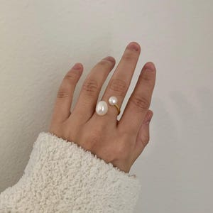 K&ouml;nnte beinhalten: Ein goldfarbener Ring mit zwei wei&szlig;en Perlen. Eine Perle ist gro&szlig; und oval, die andere kleiner und rund. Der Ring befindet sich an einer Hand, vor wei&szlig;em Hintergrund.