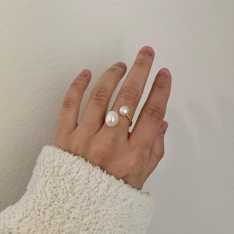 K&ouml;nnte beinhalten: Ein goldfarbener Ring mit zwei wei&szlig;en Perlen. Eine Perle ist gro&szlig; und oval, die andere kleiner und rund. Der Ring befindet sich an einer Hand, vor wei&szlig;em Hintergrund.
