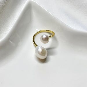 K&ouml;nnte beinhalten: Ein goldfarbener Ring mit zwei wei&szlig;en Perlen. Eine Perle ist rund, die andere oval. Das Design des Rings ist eine durchgehende Spirale, mit den Perlen an gegen&uuml;berliegenden Enden. Der Ring wird auf einer wei&szlig;en Oberfl&auml;che pr&auml;sentiert.