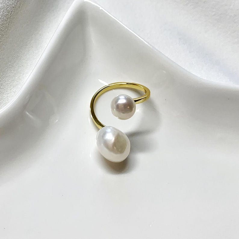 K&ouml;nnte beinhalten: Ein goldfarbener Ring mit zwei wei&szlig;en Perlen. Eine Perle ist rund, die andere oval. Das Design des Rings ist eine durchgehende Spirale, mit den Perlen an gegen&uuml;berliegenden Enden. Der Ring wird auf einer wei&szlig;en Oberfl&auml;che pr&auml;sentiert.