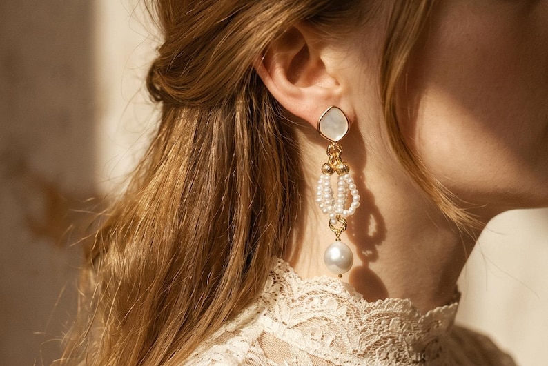 May include: A gold-toned drop earring featuring a teardrop-shaped white stone stud, a central section composed of multiple small white seed pearl loops connected by gold rings, and a single large round white pearl dangling at the very bottom.