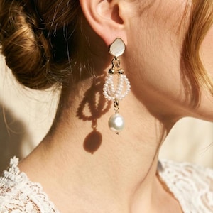 May include: Elegant gold-toned earrings with a pearlescent, irregular-shaped top, a beaded middle section, and a large, dangling pearl. The earrings are designed with a delicate, feminine aesthetic, suitable for special occasions.