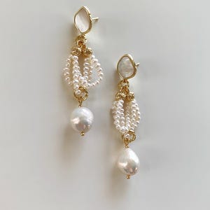 May include: A pair of drop earrings featuring a gold-tone post with a white iridescent inlay. Each earring has a cluster of small white seed pearls suspended from gold-tone metal rings, ending in a single large, irregular white baroque pearl.