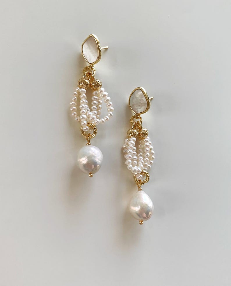 May include: A pair of drop earrings featuring a gold-tone post with a white iridescent inlay. Each earring has a cluster of small white seed pearls suspended from gold-tone metal rings, ending in a single large, irregular white baroque pearl.