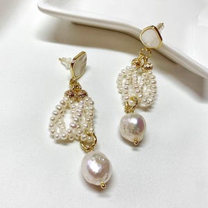 May include: A pair of gold-tone drop earrings featuring a white, irregular-shaped stone stud at the top. Below the stud, a gold-tone metal ring connects a cluster of small, white seed pearls and a single, larger, iridescent baroque pearl at the base.