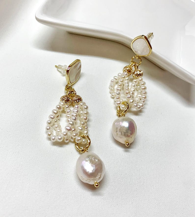 May include: A pair of gold-tone drop earrings featuring a white, irregular-shaped stone stud at the top. Below the stud, a gold-tone metal ring connects a cluster of small, white seed pearls and a single, larger, iridescent baroque pearl at the base.
