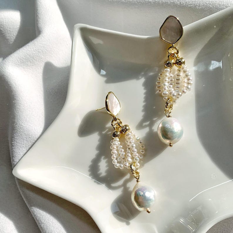 May include: A pair of gold-tone drop earrings featuring a top stud with a smooth, iridescent white inlay. Below the stud, multiple strands of tiny white seed pearls are gathered, ending in a single, large, lustrous baroque pearl pendant.