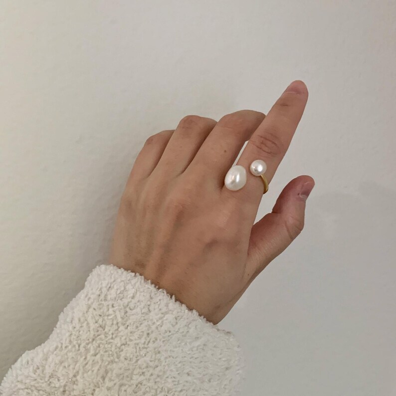 K&ouml;nnte beinhalten: Ein goldfarbener Ring mit zwei wei&szlig;en Perlen. Eine Perle ist gro&szlig; und oval, die andere kleiner und rund. Der Ring wird an einem Finger vor wei&szlig;em Hintergrund getragen.