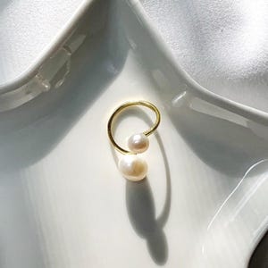 K&ouml;nnte beinhalten: Ein goldfarbener Ring mit zwei wei&szlig;en Perlen. Der Ring hat ein schlichtes, elegantes Design mit einem gebogenen Band, das die Perlen h&auml;lt. Die Perlen sind unterschiedlich gro&szlig;.