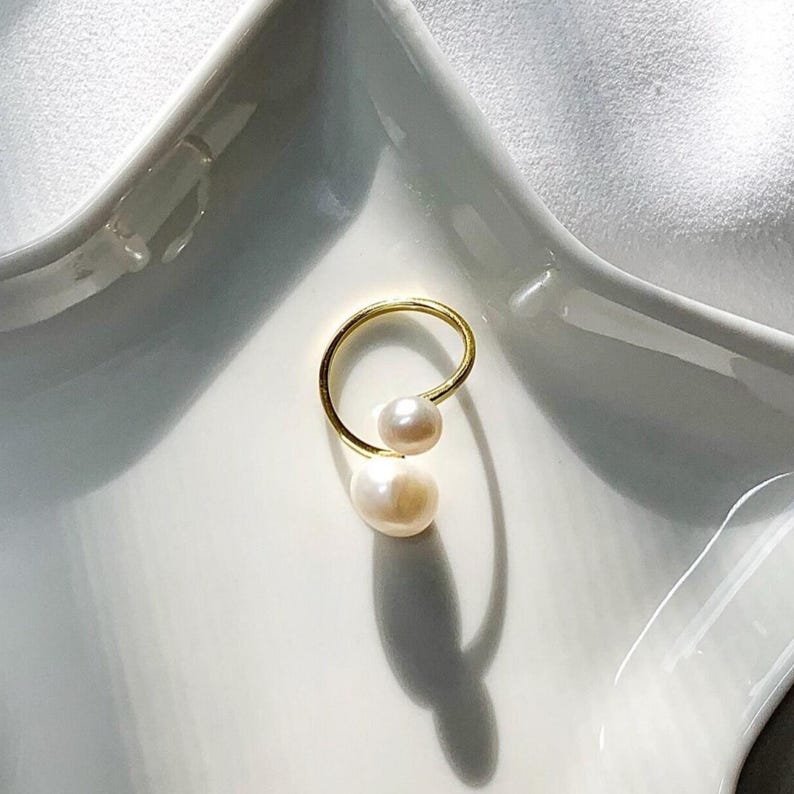 K&ouml;nnte beinhalten: Ein goldfarbener Ring mit zwei wei&szlig;en Perlen. Der Ring hat ein schlichtes, elegantes Design mit einem gebogenen Band, das die Perlen h&auml;lt. Die Perlen sind unterschiedlich gro&szlig;.