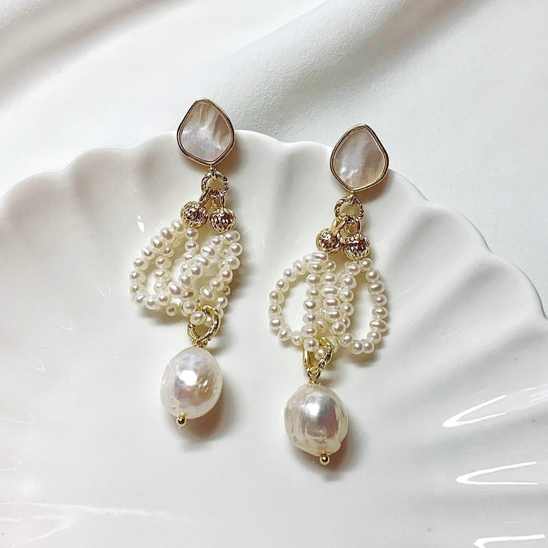 May include: A pair of drop earrings featuring a teardrop-shaped mother-of-pearl stud, gold-toned metal accents, loops of small white seed pearls, and a single larger irregular white baroque pearl dangling at the bottom of each earring.