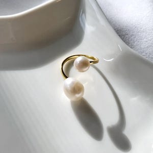 K&ouml;nnte beinhalten: Ein goldfarbener Ring mit zwei wei&szlig;en Perlen. Der Ring hat ein geschwungenes Design, mit einer Perle oben und einer gr&ouml;&szlig;eren, tropfenf&ouml;rmigen Perle unten. Der Ring ist vor einem wei&szlig;en Hintergrund platziert.
