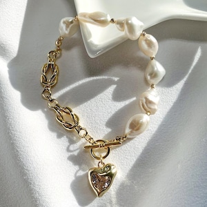 May include: A gold-toned bracelet featuring a combination of large, irregular white pearls and a decorative knot design. The bracelet includes a heart-shaped charm with a crystal center and a toggle clasp.