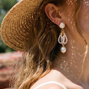 May include: A gold-tone drop earring featuring a teardrop-shaped white stone stud, a central cluster of small white seed pearls arranged in a cage-like structure, and a single large, round, off-white baroque pearl dangling at the very bottom.