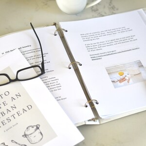 May include: A white binder with a gold ring mechanism holds pages with text and a photo of laundry drying on a clothesline. The binder is open to a page with the title "How to Create an Urban Homestead" and a drawing of a pot and a spoon. A pair of black reading glasses and two black pens are on the table.
