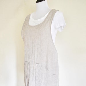 Pode incluir: Um avental de linho bege com dois bolsos grandes. O avental é sem mangas e tem um decote redondo.