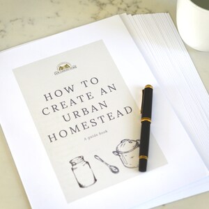 May include: A white guide book with the title "How to Create an Urban Homestead" and a guide book logo. The book is on a white surface with a black pen and a white mug.