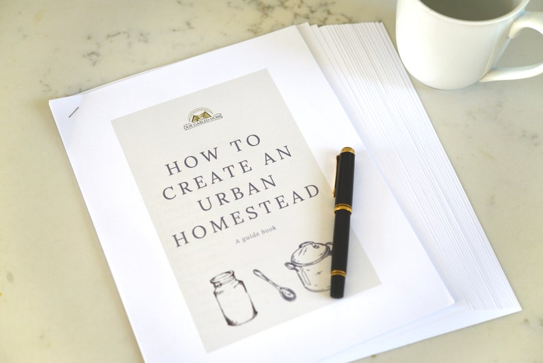 May include: A white guide book with the title "How to Create an Urban Homestead" and a guide book logo. The book is on a white surface with a black pen and a white mug.