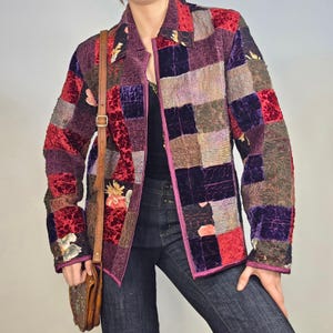 May include: A long-sleeve jacket featuring a patchwork design of various textured fabrics in shades of deep red, purple, and black. The jacket includes a collar, a front zipper closure, and scattered floral patterns. A brown leather bag strap is visible.