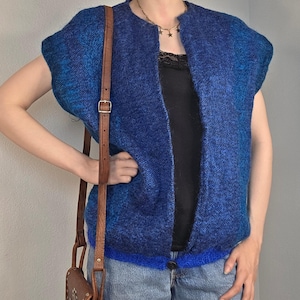 May include: A blue knit vest with short sleeves. The vest has a button closure and a gradient effect, transitioning from a darker blue to a lighter shade. A brown leather crossbody bag is visible, adding a contrasting color.