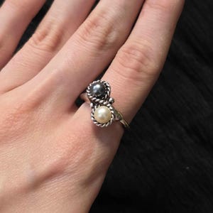 May include: A silver ring featuring two pearls, one dark gray and one cream-colored. The pearls are set in a decorative, twisted metal design. The ring is on a finger.