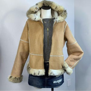 May include: A tan faux suede jacket with a faux fur-lined hood and trim. The jacket has a zip-up front and white trim accents. The jacket is displayed on a mannequin with a gray sweater and denim skirt.