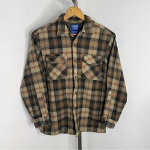 May include: A long-sleeved, button-down flannel shirt in a plaid pattern of brown, tan, and black. The shirt has two chest pockets and a collar. A small blue tag is visible near the collar.