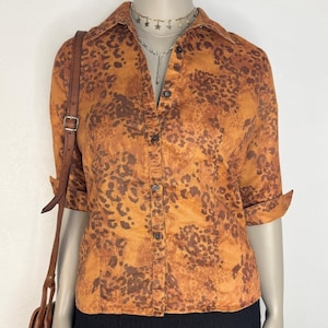 May include: An orange and brown button-down shirt with a leopard print pattern. The shirt has three-quarter sleeves and a collared neckline. The shirt is paired with a brown leather shoulder bag and a silver necklace.