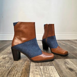 May include: A pair of ankle boots featuring a two-tone design with smooth tan leather panels and blue denim fabric sections. The boots have a square toe, a block heel, and a side zipper closure, combining leather and textile materials in one shoe.