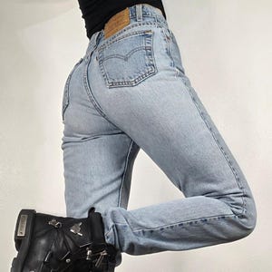 May include: Light blue denim jeans featuring a high-waisted fit, a tan leather patch on the back waistband, and a classic five-pocket design with a stitched arcuate pattern on the back pockets. The jeans are paired with a black leather boot.