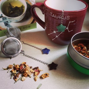 May include: A red ceramic mug with the word "Taurus" printed on it. The mug has a green star-shaped tea infuser with a chain attached. The mug is sitting on a white surface with a small tin of loose leaf tea and two tea infusers.