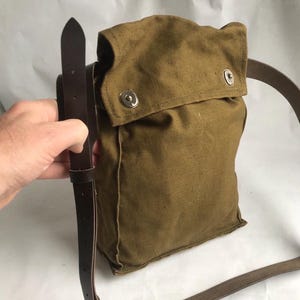Puede incluir: Una bolsa de lona verde oliva vintage con cierre de solapa asegurado por dos broches metálicos. Cuenta con una correa de hombro de cuero marrón y una correa de cuero a juego para el cierre de la bolsa. La bolsa está sujeta por una mano.
