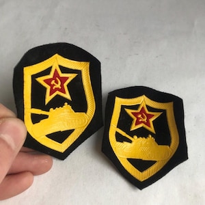 May include: Two vintage military patches. Each patch features a yellow tank and a red star with a hammer and sickle emblem on a black shield with a yellow border. The patches are likely from the Soviet era.