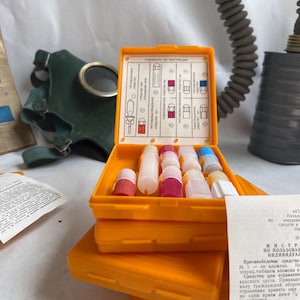 May include: An open orange first aid kit containing various vials and containers. A green gas mask and a grey filter are in the background. Several instruction sheets are also visible.