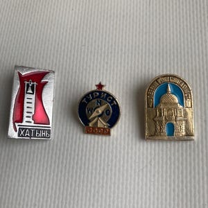 May include: Three vintage metal pins. One rectangular pin features a red and white tower with the Cyrillic word "Khatyn". A round pin has a compass design with Cyrillic text. The third pin depicts a gold building with a blue dome.