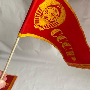 May include: A red flag with a yellow emblem and the letters "CCCP" in yellow. The flag is on a white cord and has a yellow border. A smaller version of the flag is visible in the background.