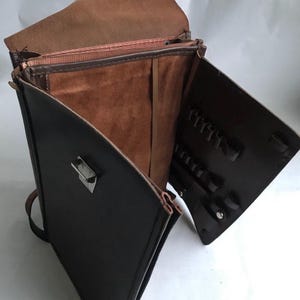 Könnte beinhalten: Eine offene, vintage Leder-Aktentasche mit braunem Innenraum und schwarzem Äußeren. Die Aktentasche hat mehrere Fächer, einen Metallverschluss und einen Schulterriemen. Der Innenraum hat eine Wildleder-ähnliche Textur und kleine Schlaufen.