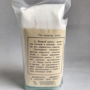 Op de afbeelding: Een verzegelde, doorzichtige plastic verpakking met een wit gaaskussen en een bruin label met Cyrillische tekst. Het label bevat instructies en een waarschuwing over de vloeistof binnenin. De tekst geeft het jaar 1981 aan.