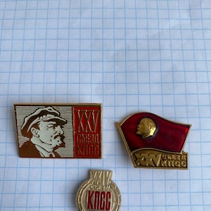 Può includere: Tre spille vintage con ritratti e testo cirillico. Una spilla rettangolare mostra il profilo di un uomo con un berretto. Un'altra spilla raffigura una bandiera rossa con un profilo. La terza spilla è rotonda con testo cirillico.