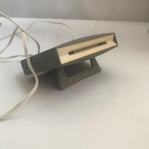 Puede incluir: Una lámpara de escritorio gris, rectangular, con interior blanco y base a juego. La lámpara tiene un diseño largo y estrecho con un cable blanco que se extiende desde la parte trasera.