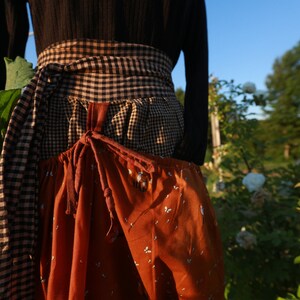 Könnte beinhalten: Eine braune und schwarze karierte Schürze mit einer großen Tasche auf der Vorderseite. Die Schürze ist mit einem braunen Band in der Taille gebunden.