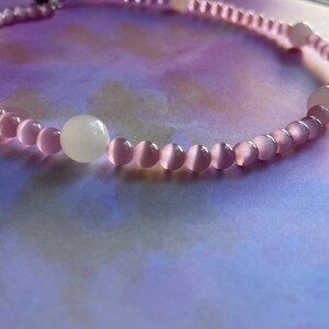 May include: A pink and white beaded necklace with a cat's eye effect. The necklace is made of small, round beads and has a simple design.