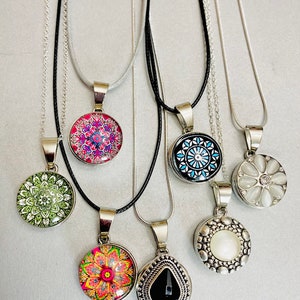 May include: Seven necklaces with different pendants. The pendants are round, with various colorful designs and patterns. Some pendants are silver with a pearl or white stone. The necklaces are on a white background.