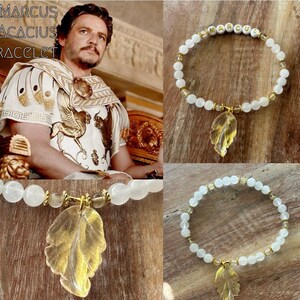 May include: White beaded bracelet with a gold leaf charm. The bracelet has the name "MARCUS" spelled out in gold letters on the beads.