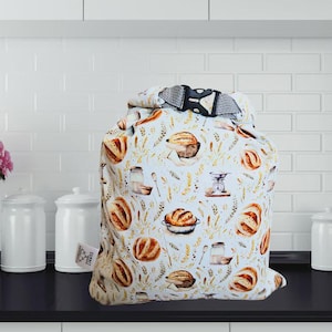 May include: A white bread storage bag with a repeating pattern of baked bread loaves, wheat stalks, and kitchen utensils. The bag has a black and gray buckle closure at the top. The bag is sitting on a black countertop.